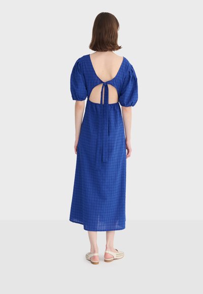 Femme portant une robe midi bleue à manches bouffantes, avec une découpe dans le dos et un détail à nouer, associée à des chaussures blanches ouvertes à l'arrière, debout de dos.