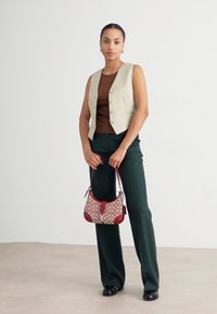 Gilet beige sopra una maglietta marrone, pantaloni larghi verdi, con in mano una borsa a tracolla rossa e marrone con dettagli rossi. Texture lisce.
