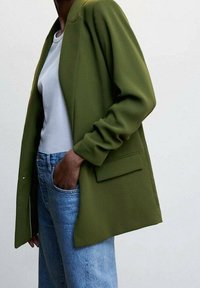 Blazer vert olive à coupe décontractée, avec une poche unique et des revers crantés, porté sur un t-shirt blanc et un jean en denim clair.