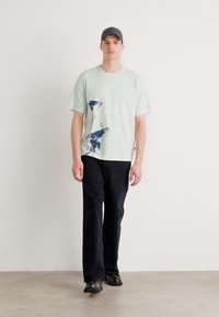Mintgrønn grafisk T-skjorte med abstrakt blått design, kombinert med mørke denimbukser og svarte sko, stående mot en nøytral bakgrunn.