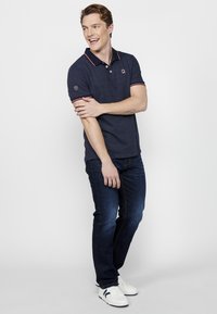 Námornícky polo tričko s červenými akcentmi a krátkymi rukávmi, skombinované s tmavými džínsami a bielymi teniskami, model stojí so založenými rukami.