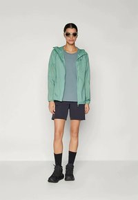 Lightweight green windbreaker with hood, paired with a gray top and black shorts. Black shoes and sunglasses complete the outdoor look.