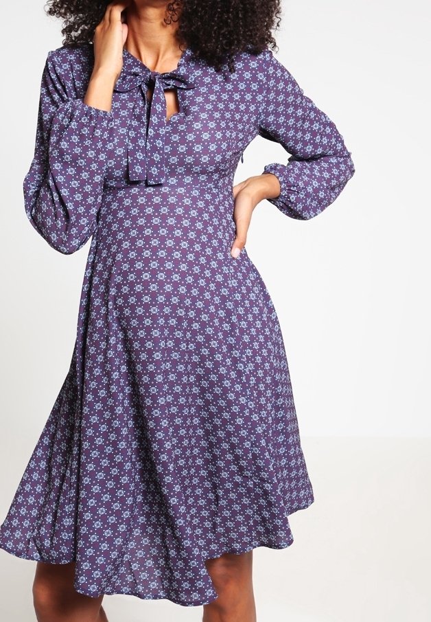 Lila blommig klänning med långa ärmar, en knuten halsringning och en utsvängd kjol. Tyget har ett litet, upprepande blommönster.