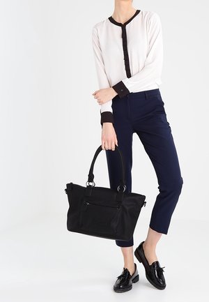 Persoon draagt witte blouse met zwarte bies, marineblauwe cropped broek, zwarte loafers, houdt grote zwarte handtas vast bij handvatten.