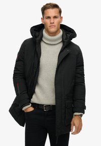 Parka aislante negra con capucha, que cuenta con bolsillos frontales, puños acanalados y detalle del logo en rojo. Debajo, un suéter de punto grueso en gris claro.