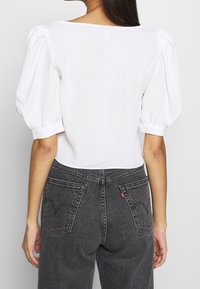 Bílá blůza s nabíranými rukávy, hladkou texturou a čtvercovým výstřihem. V kombinaci s černými džíny s vysokým pasem, které mají zadní kapsy a červenou nášivku.