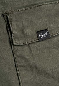 Olivgrüner Stoff mit sichtbaren Nähten und einer Tasche. Verfügt über ein schwarzes Etikett mit weißem Text "Reell est. 1997." Detail eines lässigen Kleidungsstücks.