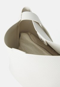 Valkoinen rei'itetty käsilaukku, jossa on leveä hihna, avoin yläosa paljastaa beige vetoketjullisen laukun sisällä valkoista taustaa vasten.