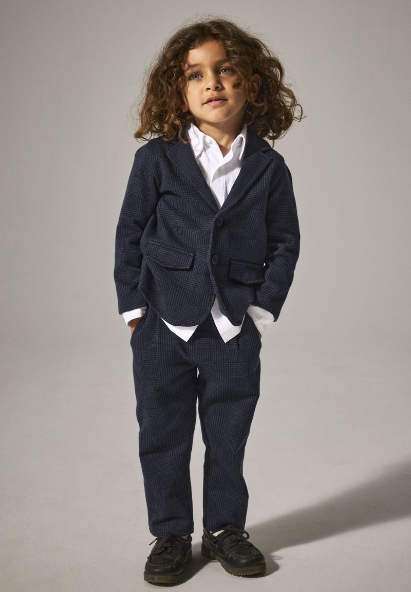 Marineblau kariertes Blazer- und Hosen-Set, weißes Hemd darunter, mit Revers und zwei Vorder Taschen, kombiniert mit schwarzen Schuhen.