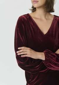 Burgunderrotes Samtkleid mit V-Ausschnitt, gefältelten Schultern und langen, gerafften Ärmeln. Glatte Textur mit drapierten Details an der Taille.