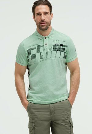 Mann mit kurzen dunklen Haaren, der ein hellgrünes Poloshirt mit schwarzem Text und olivfarbene Cargo-Shorts trägt, steht mit den Händen in den Taschen.