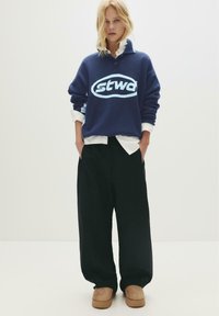 Junge Frau mit blonden Haaren, die einen marineblauen Pullover mit leicht blauem Logo, ein weißes Hemd, breite schwarze Hosen und beige Plateauschuhen trägt.