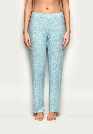 Frau trägt hellblaue Pyjamahose mit dezentem Leopardenmuster, steht barfuß vor einem schlichten weißen Hintergrund.