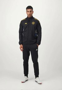 Black athletic jacket with yellow stripes and logo, paired with matching black joggers and white sports shoes. Smooth fabric, sporty design.