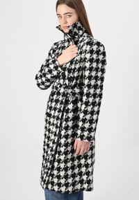 Melnā un baltā krāsā veidotā houndstooth raksta tranšejas mēteli ar augstu apkakli, pogu aizdari un piesietu jostu, izceļas ar mīkstu, teksturētu audumu.