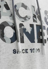Sudadera gris con letras grandes de estampado de camuflaje, cuenta con una capucha con cordones de rayas en blanco y negro. El texto incluye "DESDE 1990".