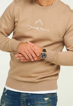 Mężczyzna ubrany w beżową bluzę Jack & Jones i zegarek na rękę z czarnym paskiem, w połączeniu z postrzępionymi niebieskimi jeansami, ręce splecione na talii.