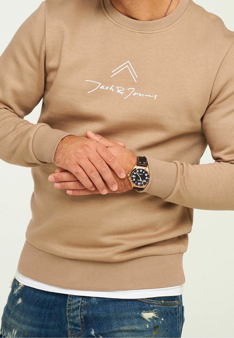 Homme portant un sweat-shirt beige Jack & Jones et une montre-bracelet avec un bracelet noir, associé à un jean bleu déchiré, les mains jointes à la taille.