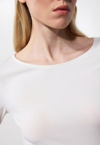 Weißes, tailliertes Langarmshirt mit rundem Halsausschnitt, glatter Textur und dezenten Nähten, die den Kragen und die Schultern betonen.