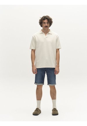 Mies, jolla kiharat hiukset ja viikset, pukeutuneena kermanväriseen poolopaitaan, käännettyihin farkkushortseihin, valkoisiin sukkiin ja ruskeisiin slip-on -kenkiin, seisoo valkoisella taustalla.