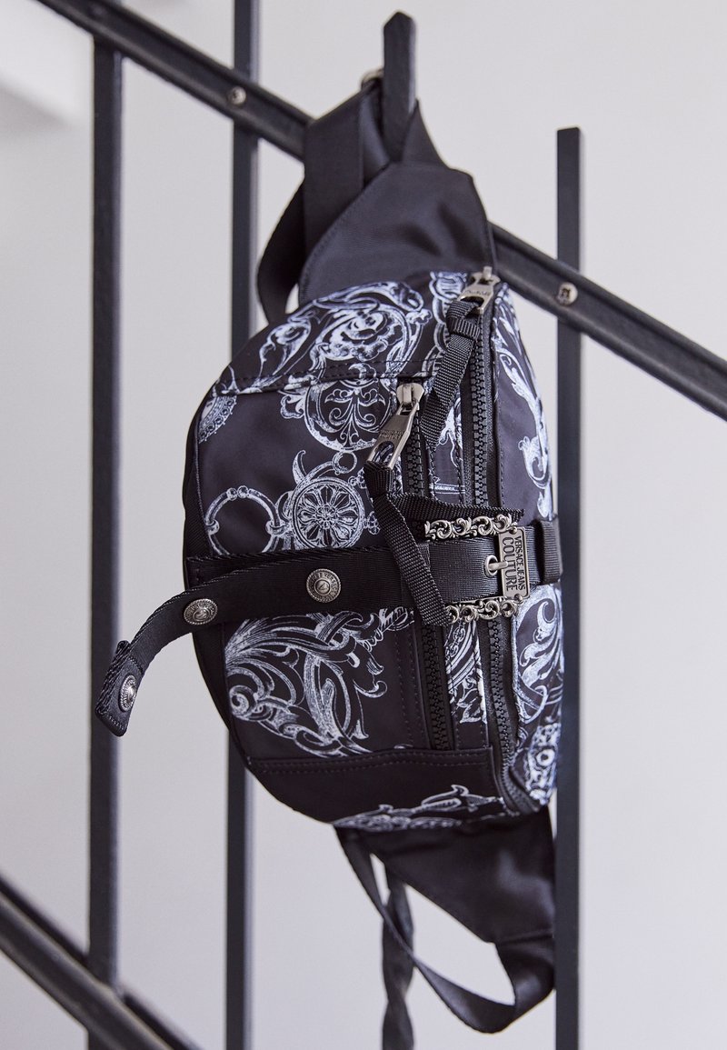 Black bag with a white floral pattern, featuring zippers and metal accents. Made from fabric with a textured look; adjustable strap.