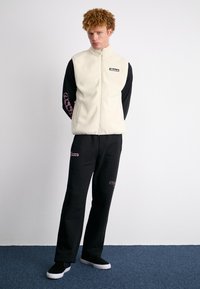 Fleeces veste gaišā krēmkrāsā ar rāvējslēdzēju; melna krekla ar garām piedurknēm un rozā grafiku uz piedurknēm; melnas bikses ar tekstu; melnas kedas.