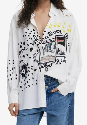 Žena v bílé košili na knoflíky s černými a žlutými abstraktními vzory, textem a fotokolážovým potiskem, v kombinaci s modrými džínami.