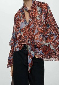 Blouse transparente à motifs floraux rouges, bleus et crème, manches longues à volants et col noué, portée avec un pantalon noir.