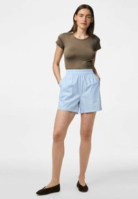 Brūns pieguļošs T-krekls, ko valkā ar gaiši ziliem svītrainiem šortiem ar elastīgu jostu un sānu kabatām, kopā ar tumšām slīdošām apaviem.