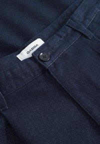 Marineblaue Denim-Hosen mit glatter Textur, versehen mit einem schwarzen Knopf, einem weißen Etikett mit "GABBA" und sauberer Naht entlang des Bundes.