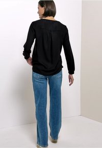 Schwarze Langarmbluse mit einem dezenten Rückenteil-Detail, kombiniert mit hellblauen Schlagjeans. Der Stoff wirkt weich und hat eine glatte Textur.