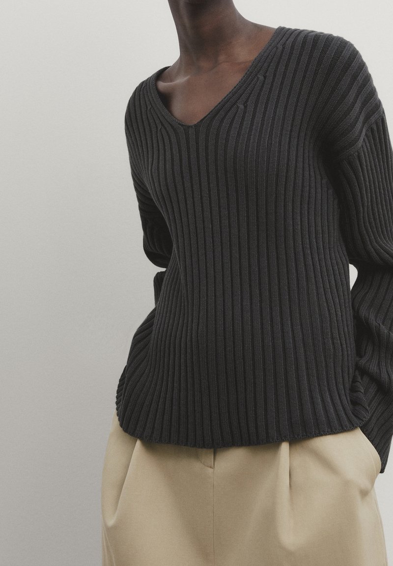 Personne portant un pull noir à col en V côtelé et un pantalon beige, les mains dans les poches, sur un fond clair uni.