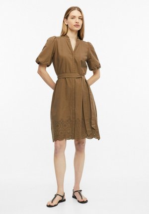 Femme debout portant une robe marron mi-genou avec manches ballon, une ceinture à la taille et un ourlet décoratif ajouré, associée à des sandales noires.