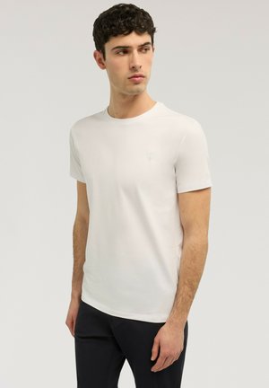 Junger Mann mit kurzen lockigen Haaren, der ein schlichtes weißes kurzärmeliges T-Shirt und dunkle Hosen trägt, blickt seitlich vor einem einfarbigen Hintergrund.