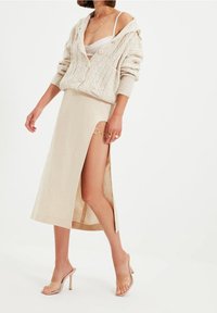 Krémový cardigan s pleteným vzorem a kapucí, doplněný béžovou sukní ke kolenům s vysokým rozparkem. Průhledné sandály na podpatku.