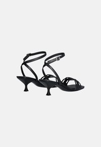 Sandali neri con tacco, design a fascia incrociata, realizzati in materiale liscio. Tacco curvato prominente e stile aperto davanti. Cinghie per caviglia regolabili.