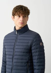 Veste en duvet marine avec un col montant, fermeture éclair et design matelassé horizontal. Présente un patch logo sur la manche supérieure. Texture lisse.