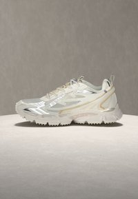 Baskets de sport beige clair et blanches avec panneaux en mesh, détails réfléchissants, semelle épaisse à crampons et languettes de tirage sur la languette et le talon.