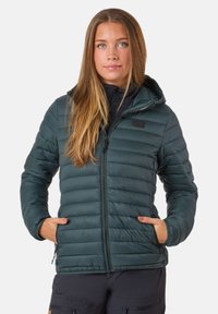 Junge Frau mit langen Haaren, die eine dunkelgrüne gesteppte Jacke mit Kapuze trägt und die Hände in den Taschen hat, steht vor einem schlichten Hintergrund.