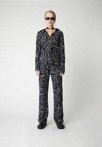 Schwarz- und graumusterter Trainingsanzug mit langen Ärmeln und bequemen Hosen, ausgestattet mit einem strukturierten Stoffdesign. Schwarze klobige Schuhe und Sonnenbrille.