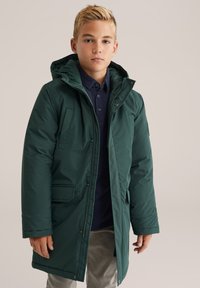 Zēns, kurš valkā tumši zaļu ziemas mēteli ar kapuci, virs zilā krekla un pelēkajiem biksēm, stāv uz vienkārša bēša fona.