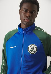 Blue and green zip-up jacket made of smooth fabric, featuring a basketball logo and Nike swoosh on the chest, with white accents.