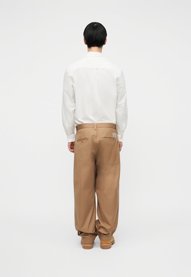 White long-sleeve shirt with a button-down collar, paired with loose brown pants featuring a back pocket and a logo tag. Neutral background.