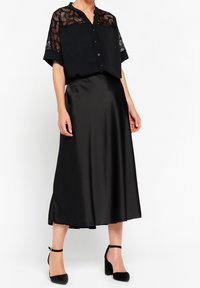 Blusa nera cropped con dettagli in pizzo sulle spalle, abbinata a una gonna midi in satin nera e décolleté nere con cinturino alla caviglia.