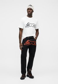 Λευκό μπλουζάκι με γραφικό μαύρης γάτας, συνδυασμένο με μαύρο παντελόνι που έχει σχέδιο πορτοκαλί γάτας. Μαύρα μποτάκια ολοκληρώνουν την εμφάνιση.