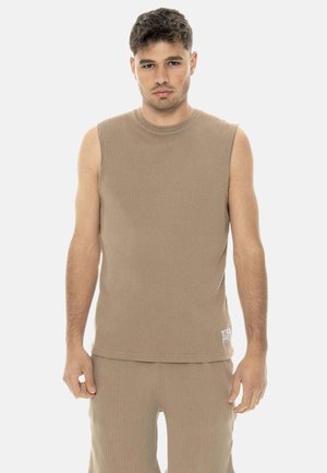 Junger Mann, der ein ärmelloses, beige geripptes Oberteil und passende gerippte Hose trägt, mit neutralem Gesichtsausdruck vor einfachem Hintergrund stehend.