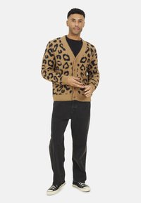 Cardigan en imprimé léopard dans les tons beige et noir, texture tricotée, fermeture à boutons, porté sur une chemise noire, associé à un jean noir et des baskets.