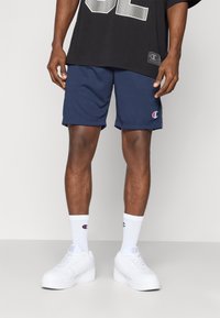 Sötétkék sportnadrág oldalsó logóval, fehér sportcipővel és zoknival párosítva. Túlnyújtott fekete póló, merész ezüst grafikával.