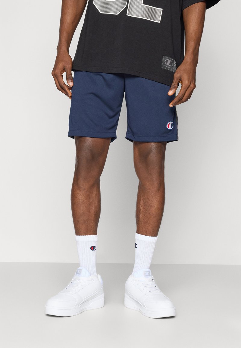 Sötétkék sportnadrág oldalsó logóval, fehér sportcipővel és zoknival párosítva. Túlnyújtott fekete póló, merész ezüst grafikával.