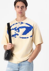Beżowy t-shirt z krótkim rękawem z dużą niebieską abstrakcyjną grafiką. Model trzyma małą czarną torbę, nosząc jasne dżinsy.